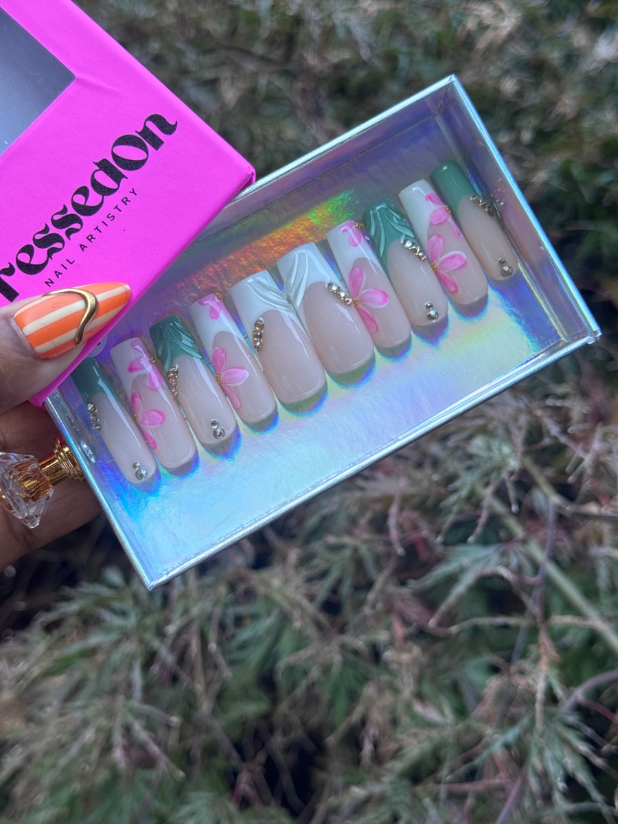 Set of medium length press-on nails with a nude base, pink flower petal art, green and white French tips, and small gems.