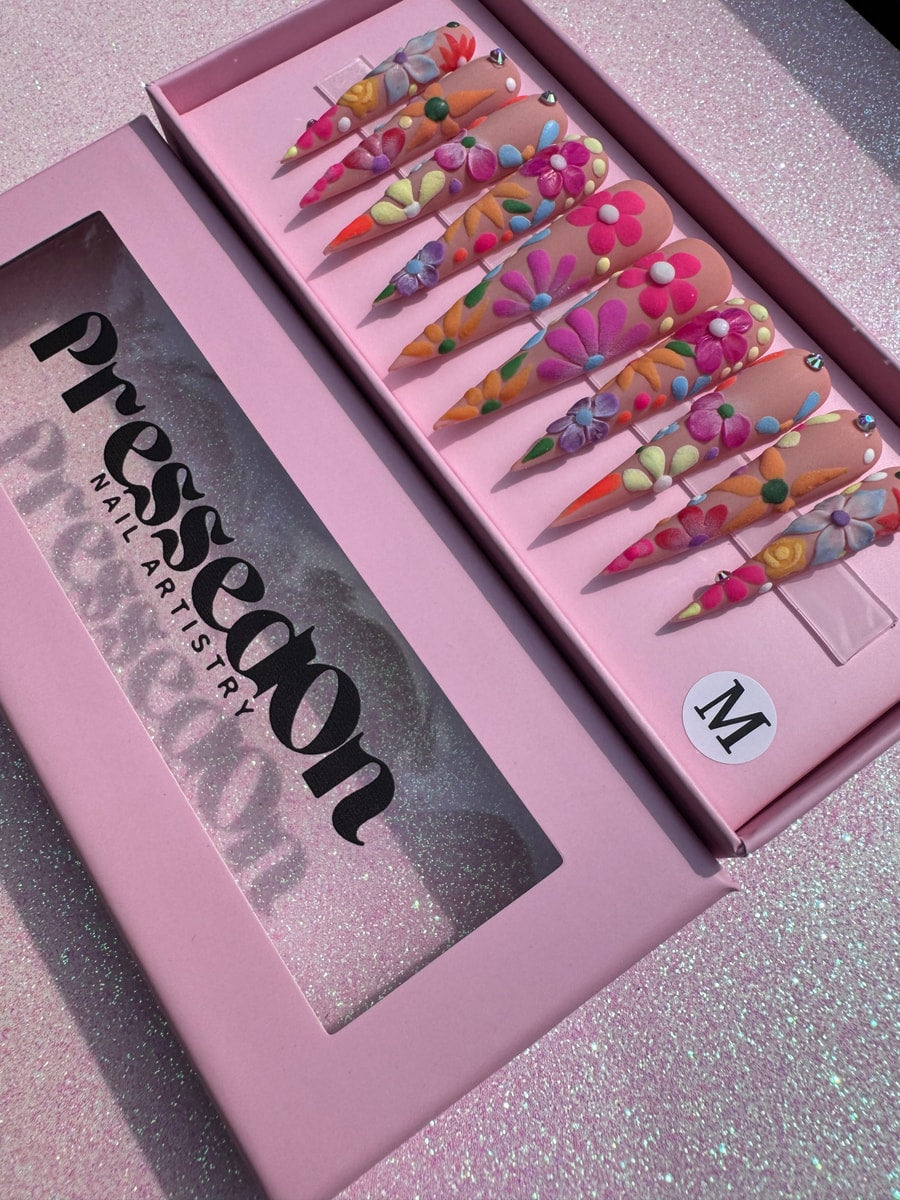 Extra long stiletto press-on nails with a colorful floral design. The nails feature flowers, dots, and gems on a nude backdrop.