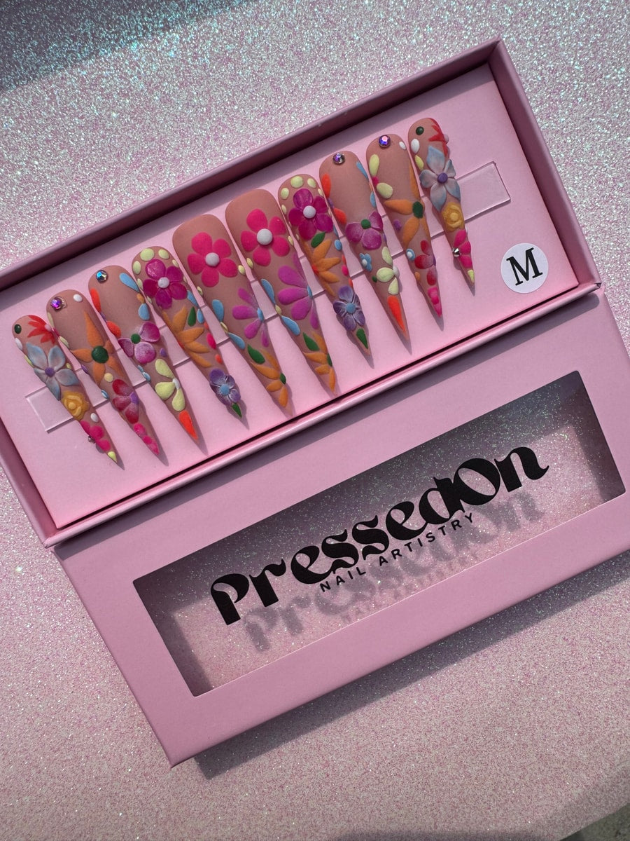 Xtra long stiletto press-on nails with a nude base and colorful 3D floral nail art. The nails are decorated with pink, orange, yellow, blue, and purple flowers and rhinestone gems.