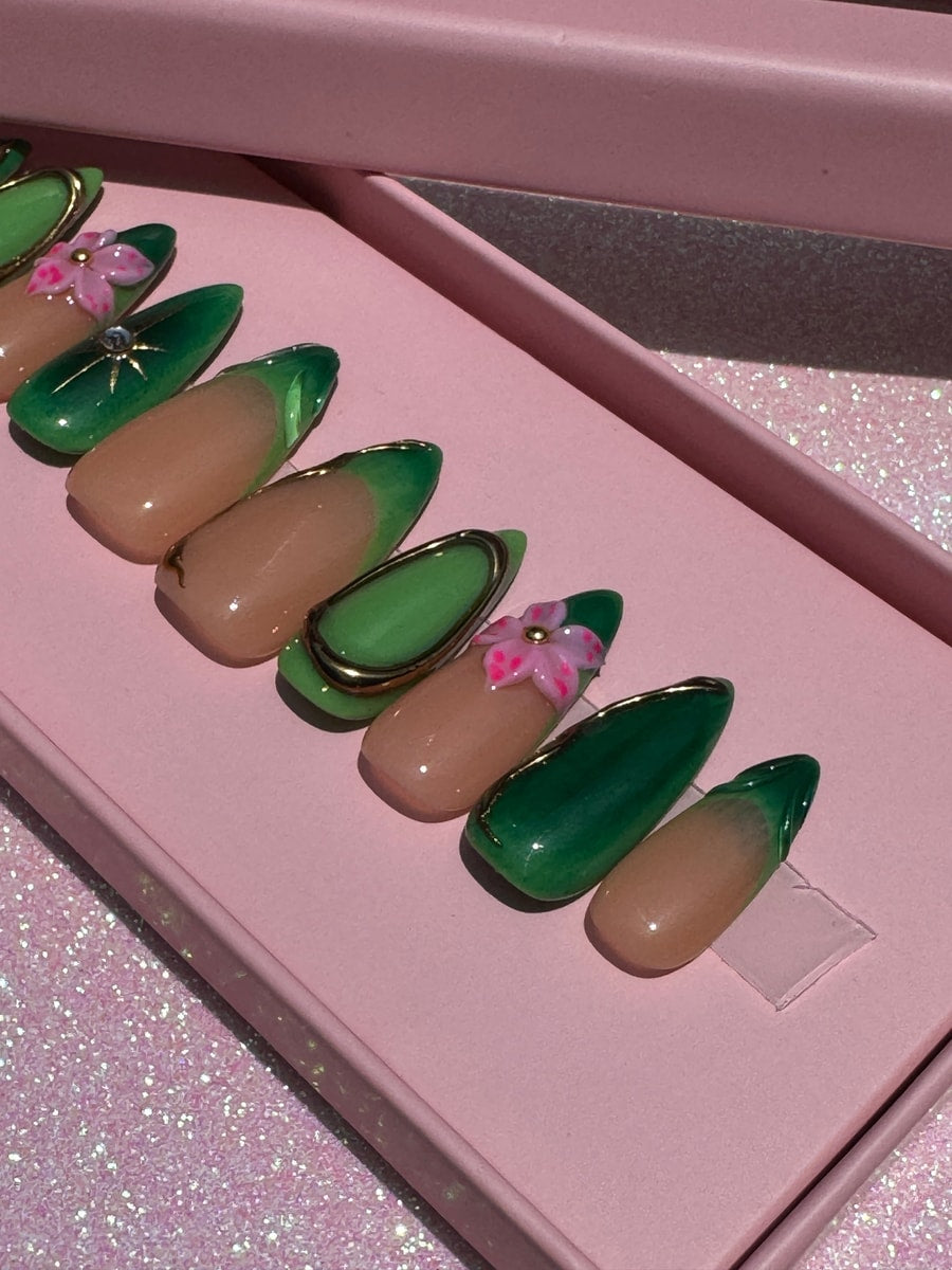 Almond shaped press-on nails with nude and green designs. Several nails feature 3D pink flower accents and gold outlines.