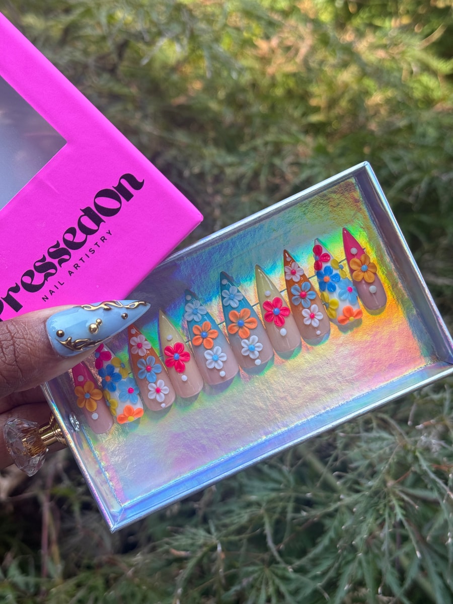 Stiletto press-on nails featuring colorful 3D flower designs. Colors include pink, orange, white, blue, and yellow on a light nude base.