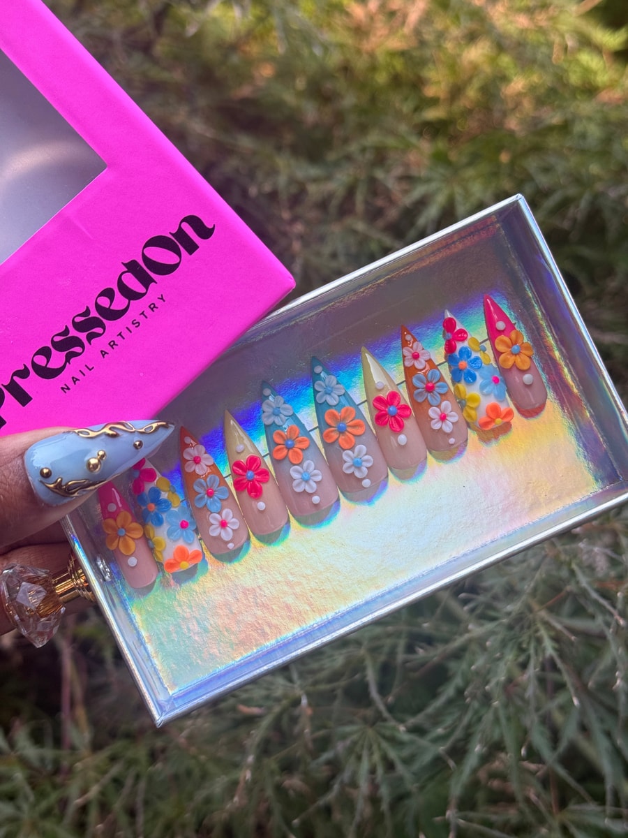 Short stiletto press-on nails with a nude base, decorated with colorful 3D flower designs in a variety of colors, including orange, blue, pink, and white.