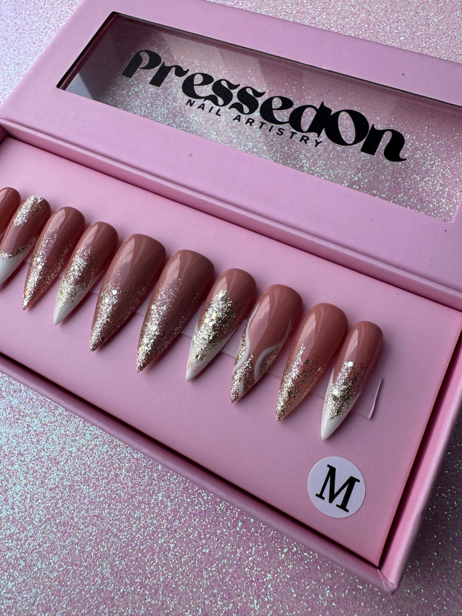 Medium length pointed press-on nails with French tips, nude base, and gold glitter accents.