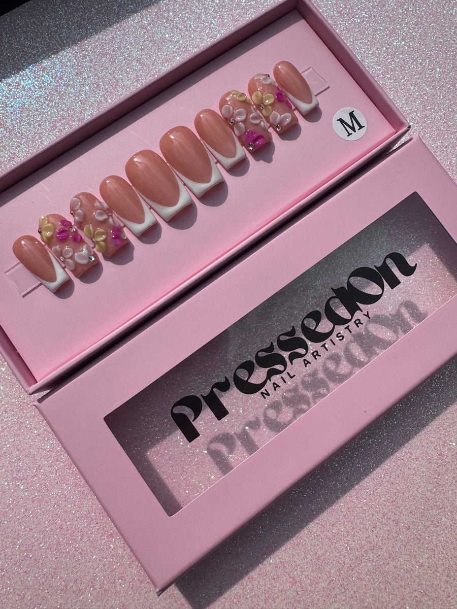 Short length press-on nails featuring a nude base with classic white French tips. A few nails have 3D floral decorations in pink, yellow, and white.