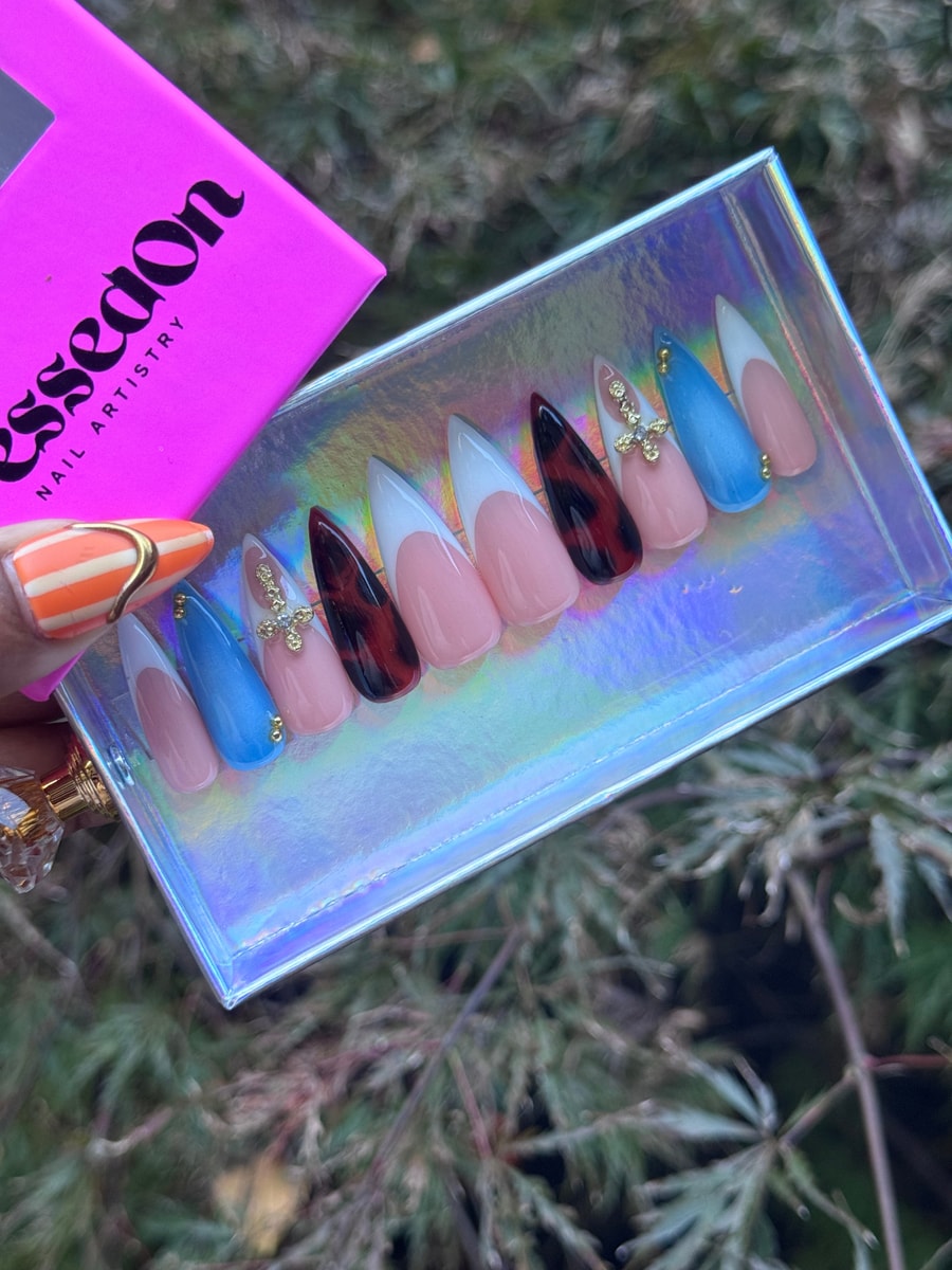 Stiletto shaped medium-length press-on nails feature French tip, solid blue, and tortoiseshell designs. Some nails have cross charms and gold bead accents.
