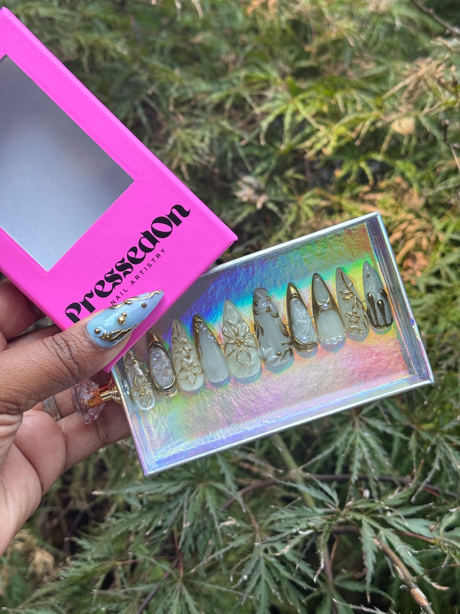 Short stiletto press-on nails feature a frosty white finish with gold floral accents and pearls. One nail has a light blue base with similar gold embellishments.