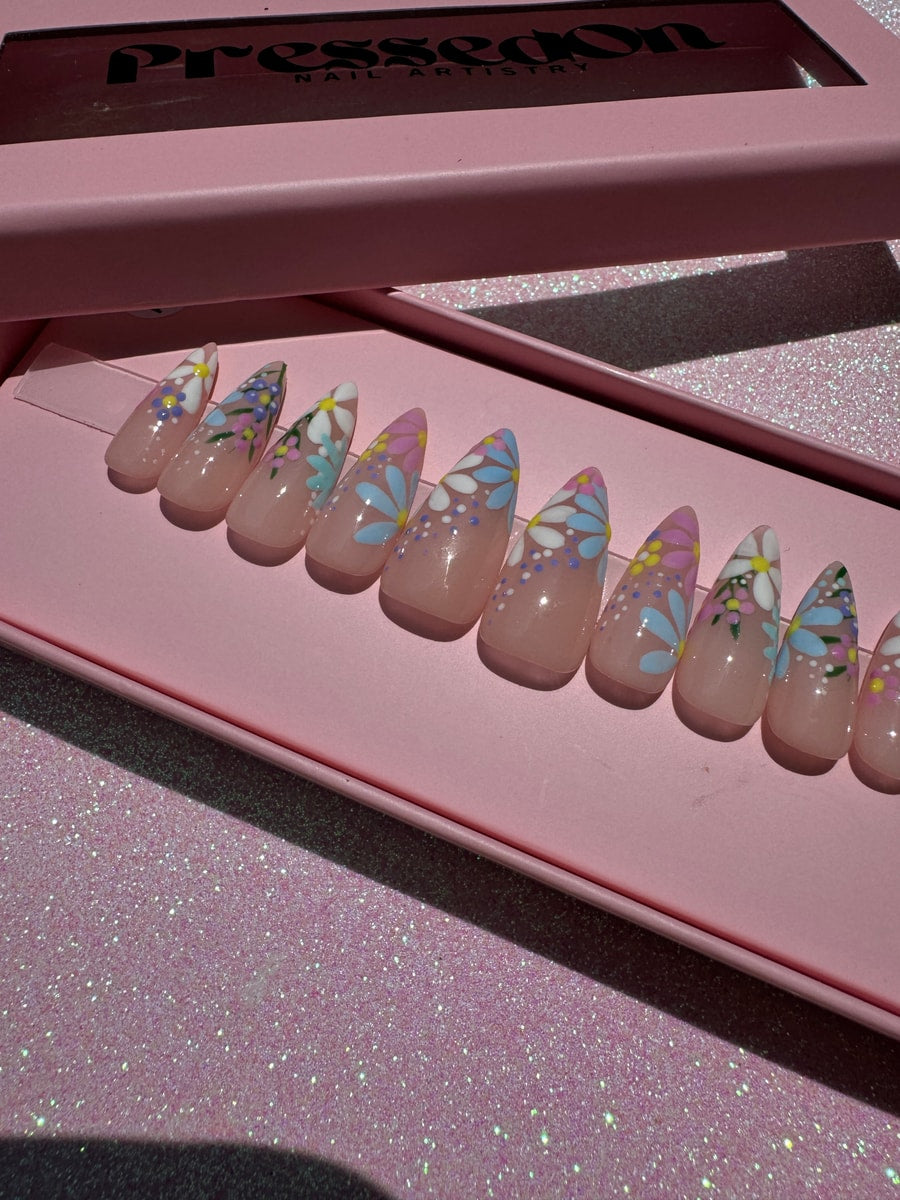 Short almond shaped press-on nails with a garden floral design. The nails have a clear base with white, pink, blue, and yellow flowers and green leaves.