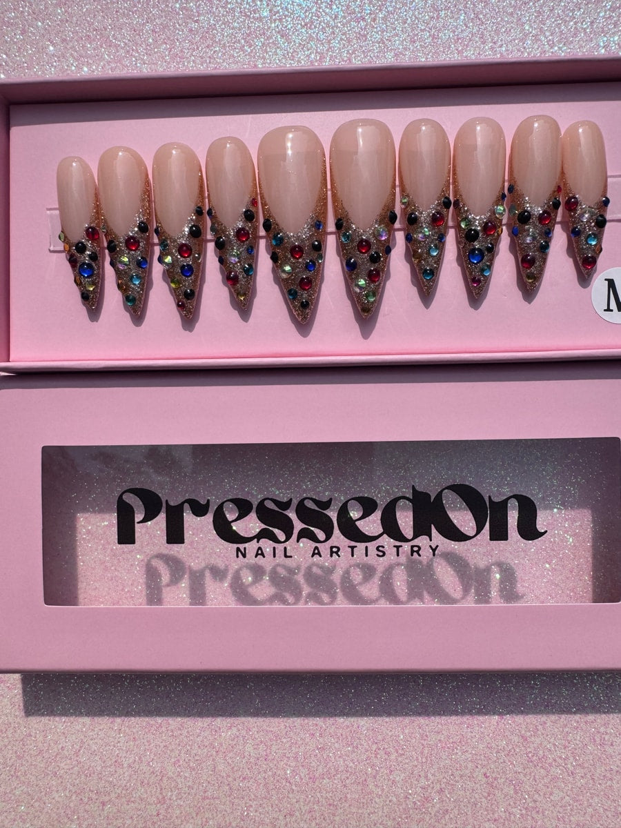 Medium length press-on nails feature gold glitter and colorful gem accents. The nails have a nude base and gem decorations near the tips.