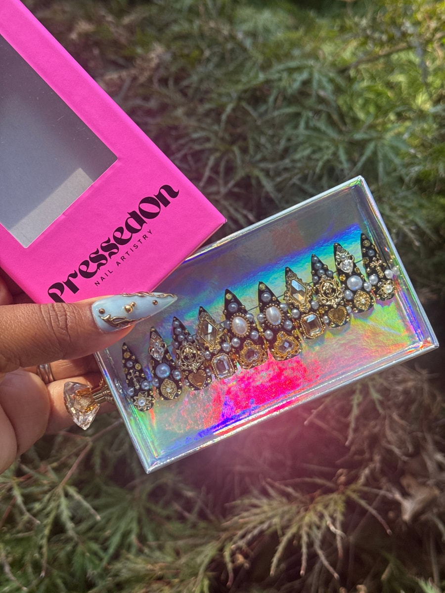 Short press-on nails with black tips and an array of gems and pearls as decoration. The nails feature silver and gold embellishments.