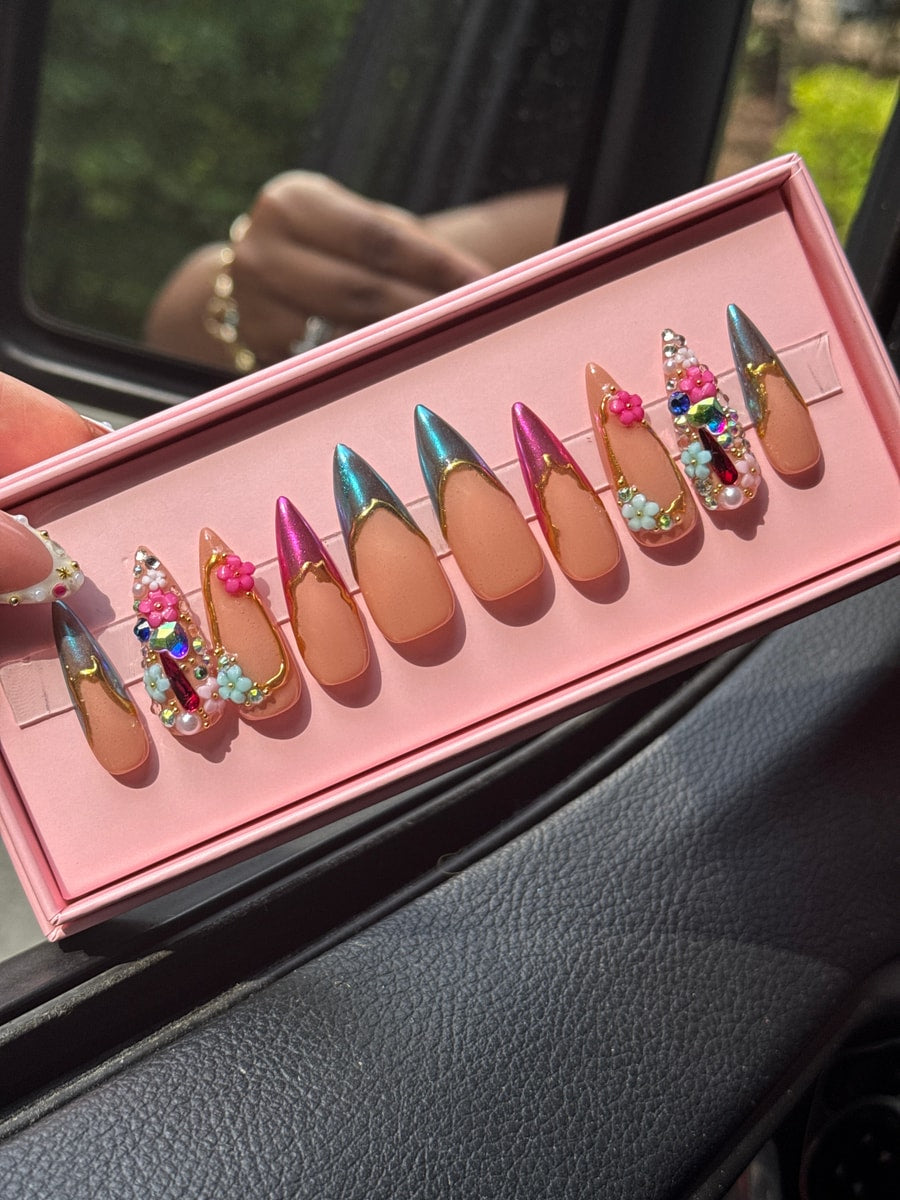 Set of stiletto shaped press-on nails with nude base and chrome accents. Some nails feature gem embellishments, floral details, and gold detailing.