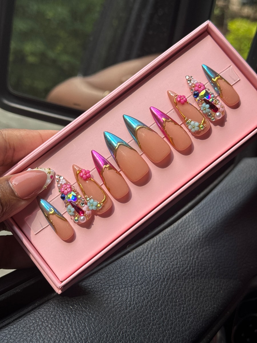 Medium-length press-on nails with nude base, iridescent chrome tips, floral details, and gem decorations.