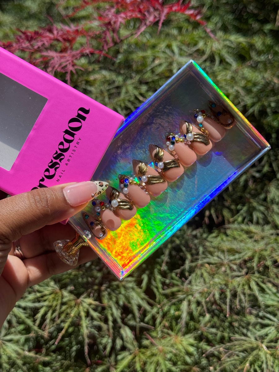 Short, nude-colored press-on nails with gold accents, pearls, and colorful gemstones. One nail has a cheetah print french tip.
