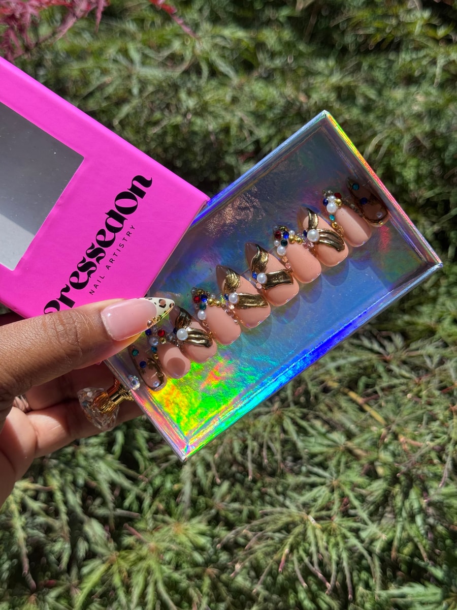 Short nude press-on nails are decorated with gold accents, pearl details, and colorful gems.