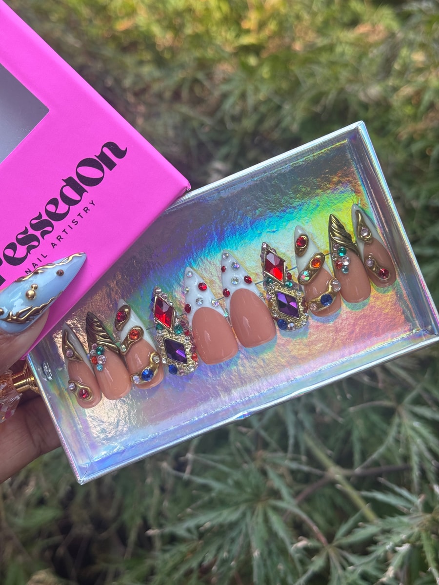 Short press-on nails featuring a nude base, white tips, and colorful gemstone embellishments. These nails are decorated with rhinestones and ornate gold accents.