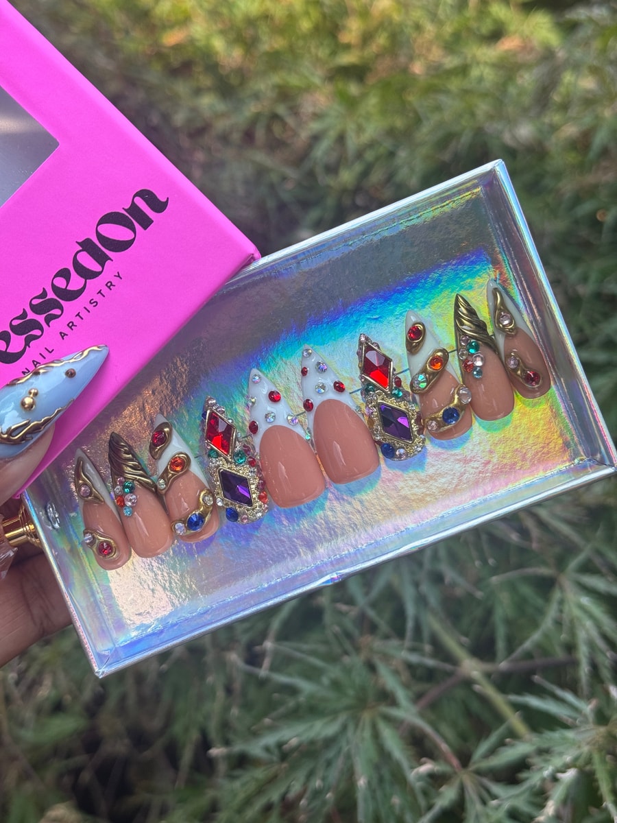 Set of short oval press-on nails with a nude base, adorned with colorful gems, rhinestones, and gold decorative elements.