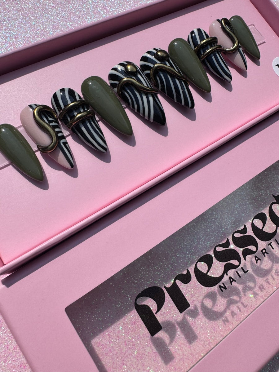 Stiletto shaped press-on nails show geometric designs with olive green and zebra stripe patterns, each accented with a gold snake-like charm.