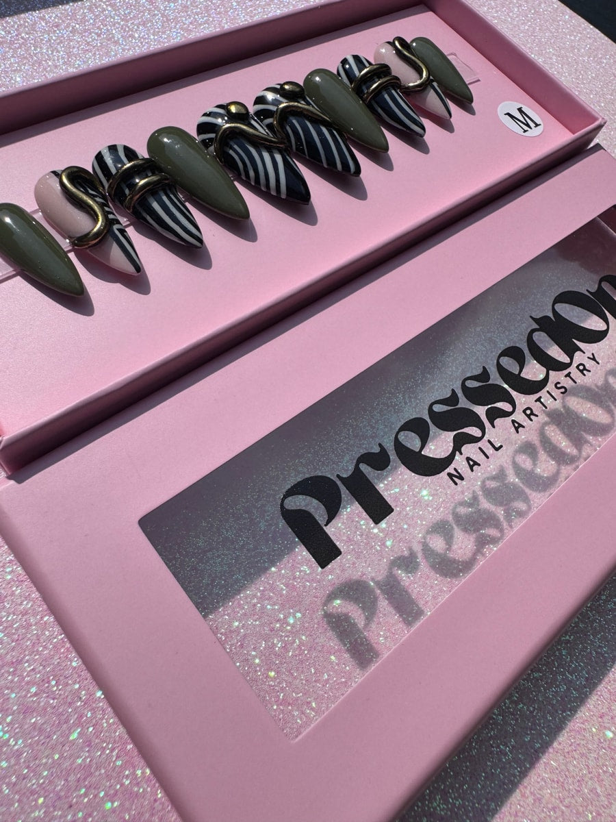 A set of super short stiletto press-on nails with geometric designs. Some nails are striped, some are solid olive green, and one is light pink, all with gold accents.