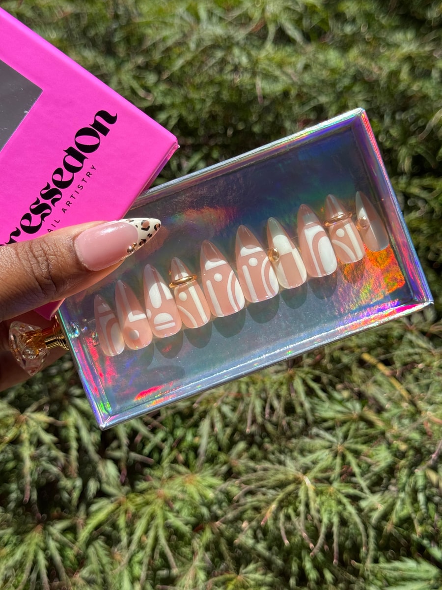 Short nude almond press-on nails feature a geometric white design. Some nails also have small gold bead accents.