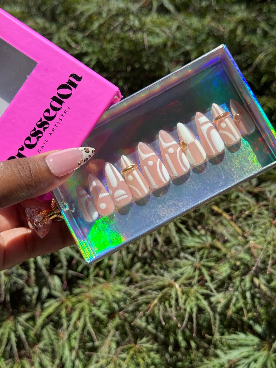 Short almond press-on nails with a tan base and white geometric designs. Several nails feature gold rings and accents.