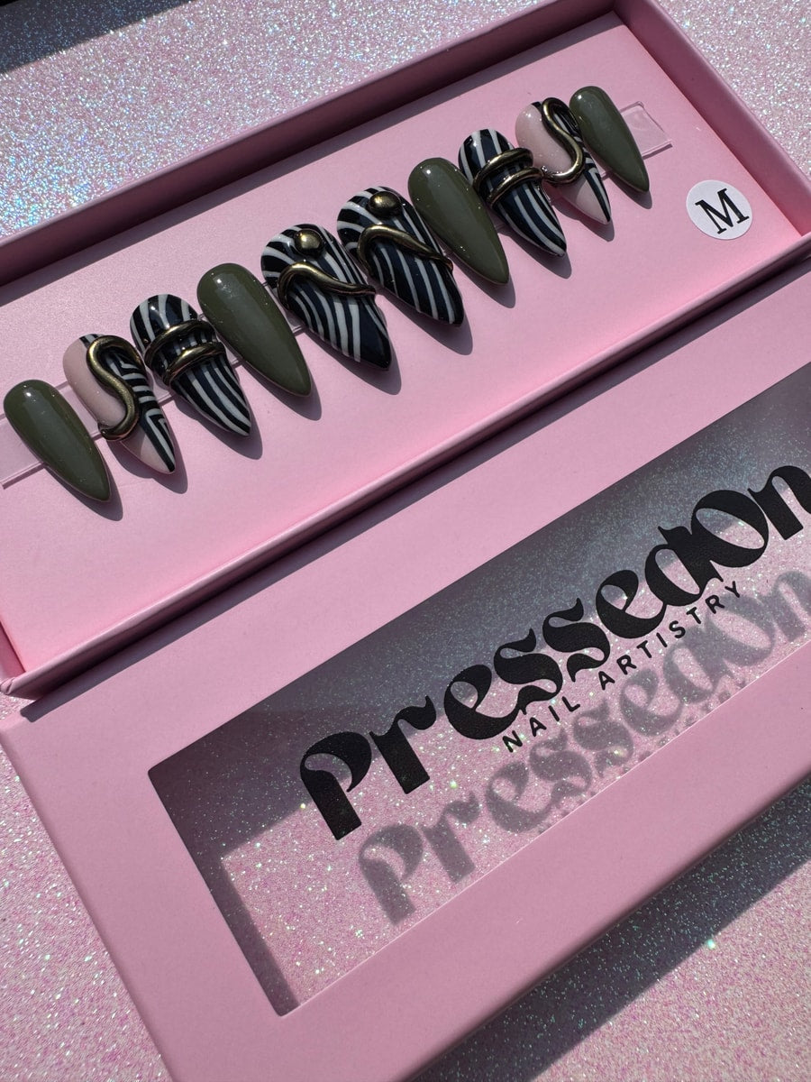 A set of short press-on nails featuring a geometric design with zebra stripes, solid olive green, and pale pink. Several nails have a decorative gold wire accent.