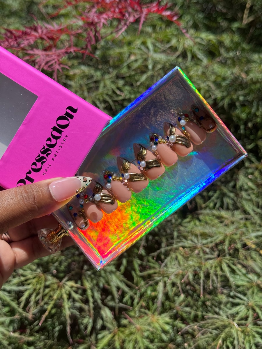 Short nude almond press-on nails are decorated with gold detailing, pearl gems, and colorful rhinestones for a glamorous look.