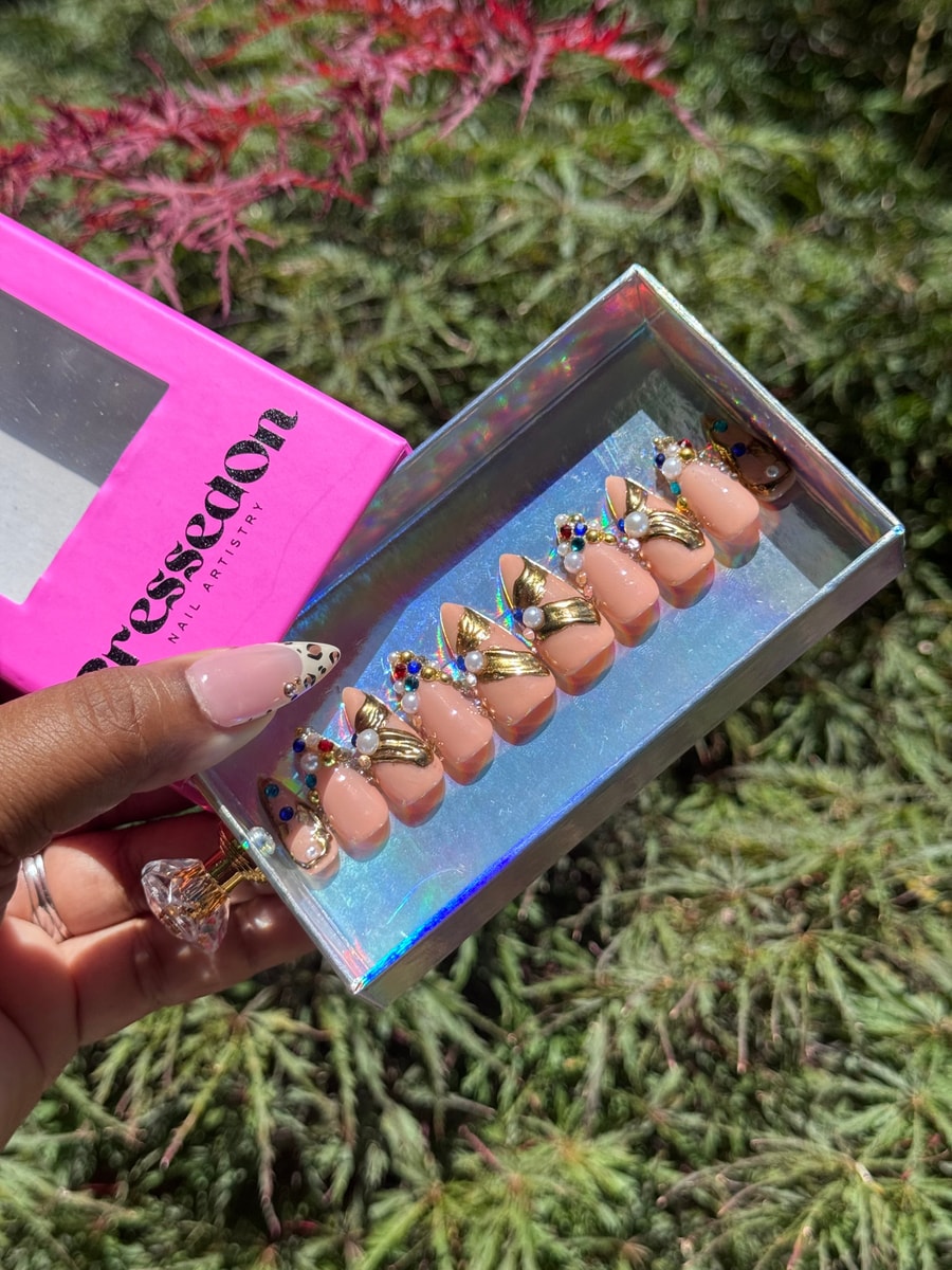 Short peach-colored press-on nails are decorated with gold accents, pearl embellishments, and colorful gemstone beads.