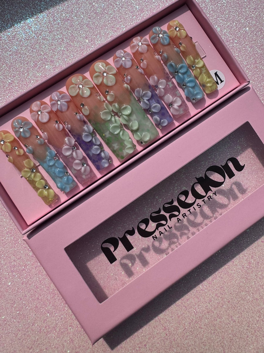 A set of long, colorful press-on nails decorated with 3D flowers and small glitz accents. Each nail has a different pastel color.