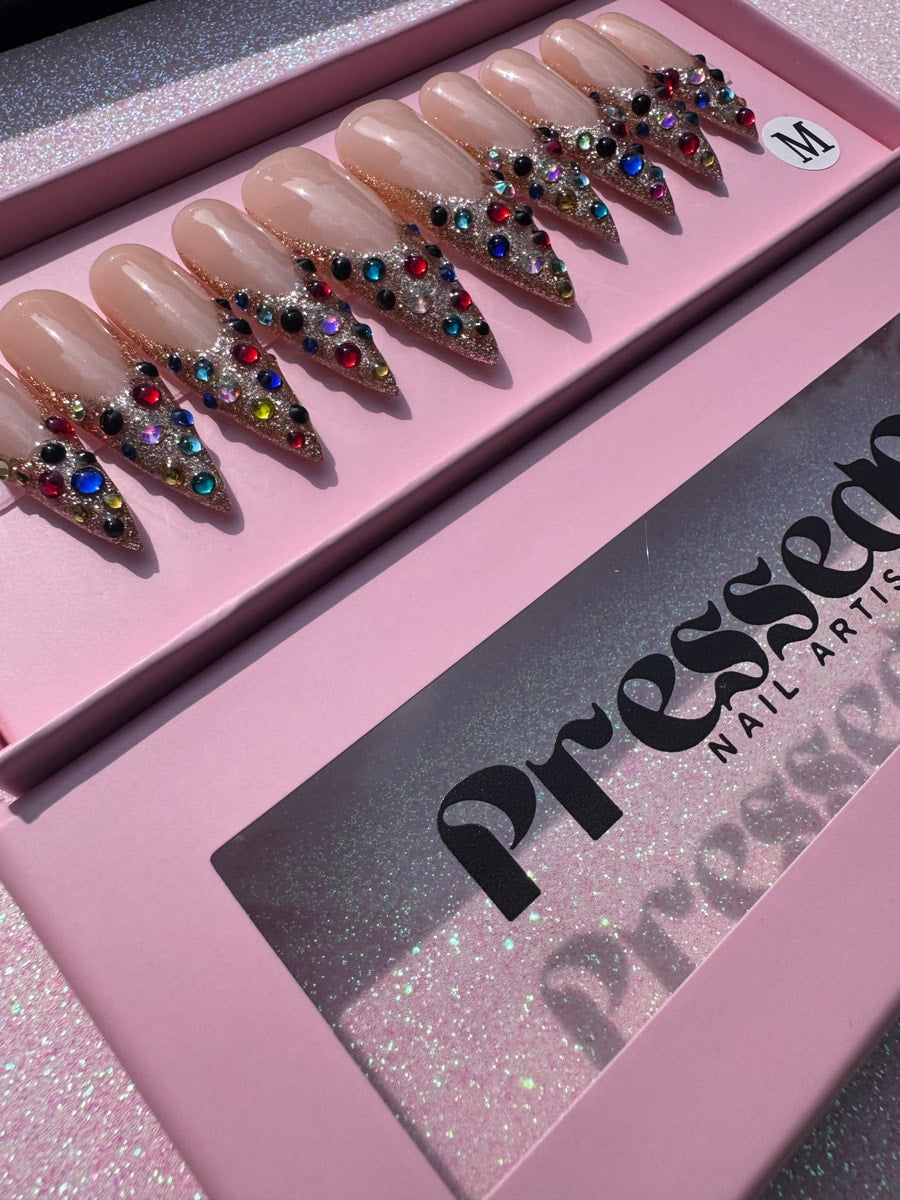 Medium press-on nails with gold glitter and colorful gems decorating the tips.