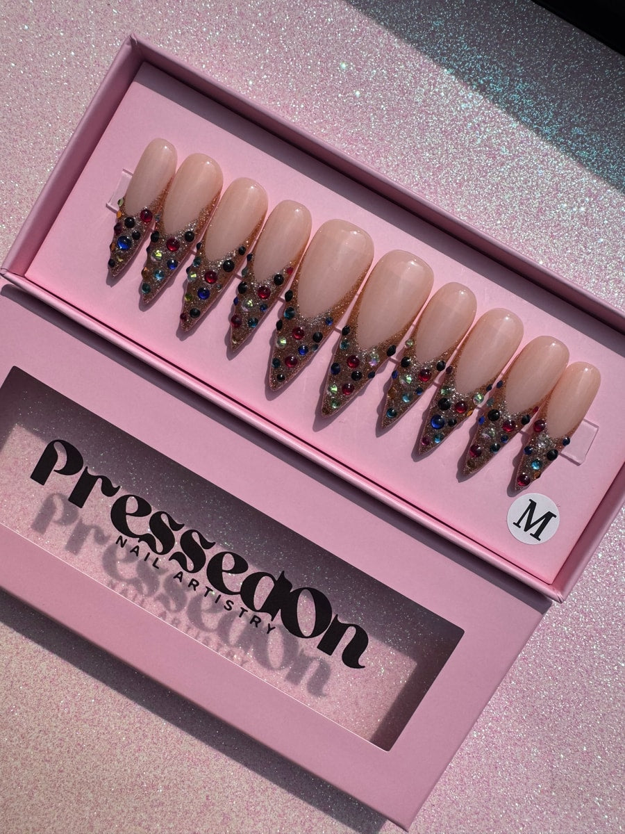 Medium-length press-on nails feature a nude base with gold glitter french tips decorated with colorful gems.