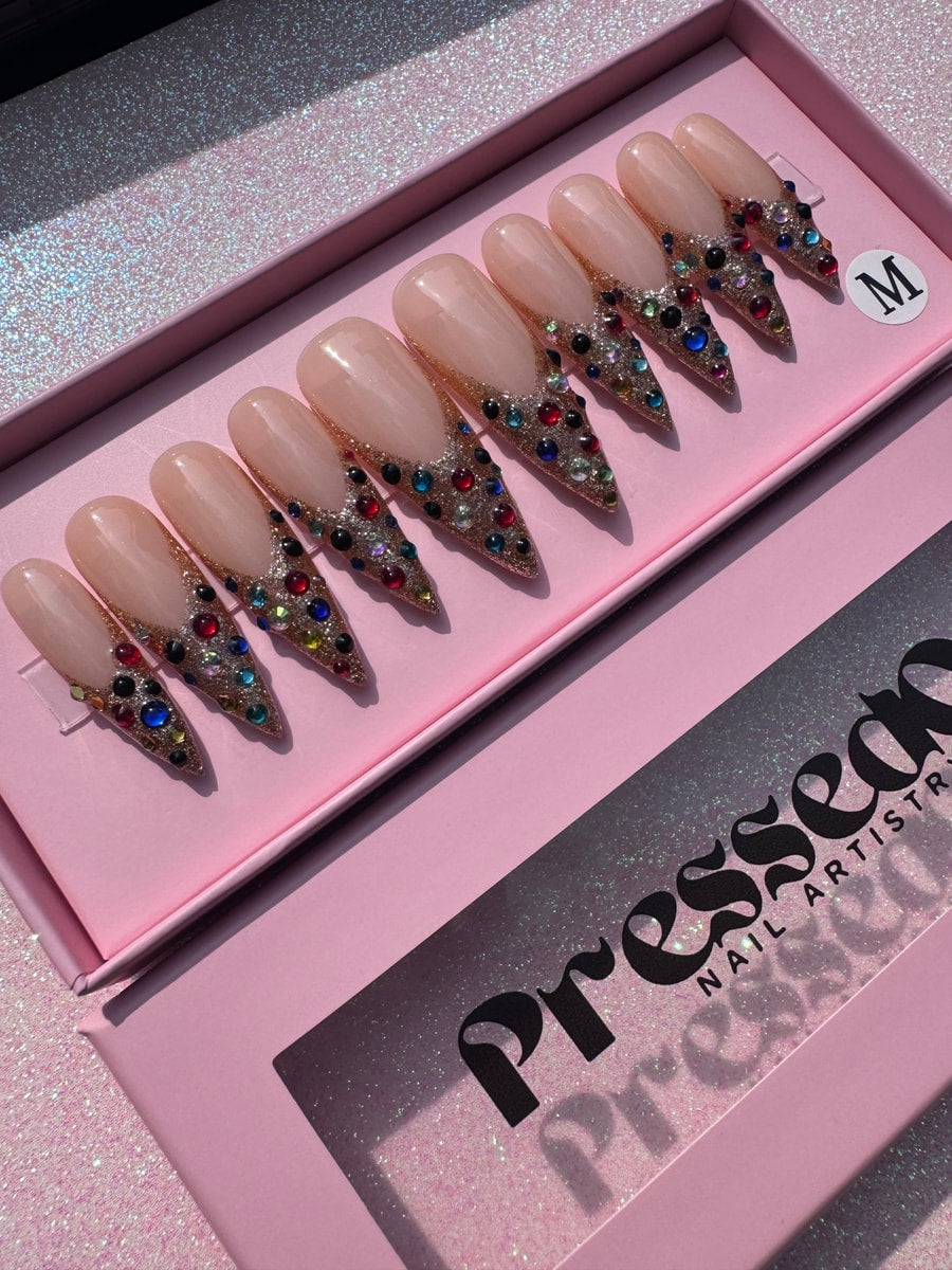 Set of medium-length press-on nails featuring a nude base, gold glitter french tips, and assorted colorful gems.