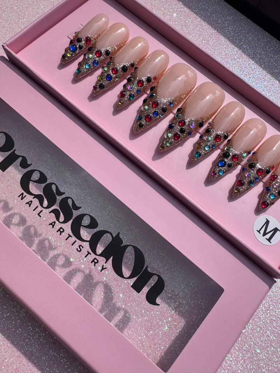 Medium press-on nails feature a nude base, gold glitter detailing, and colorful gems near the tips.