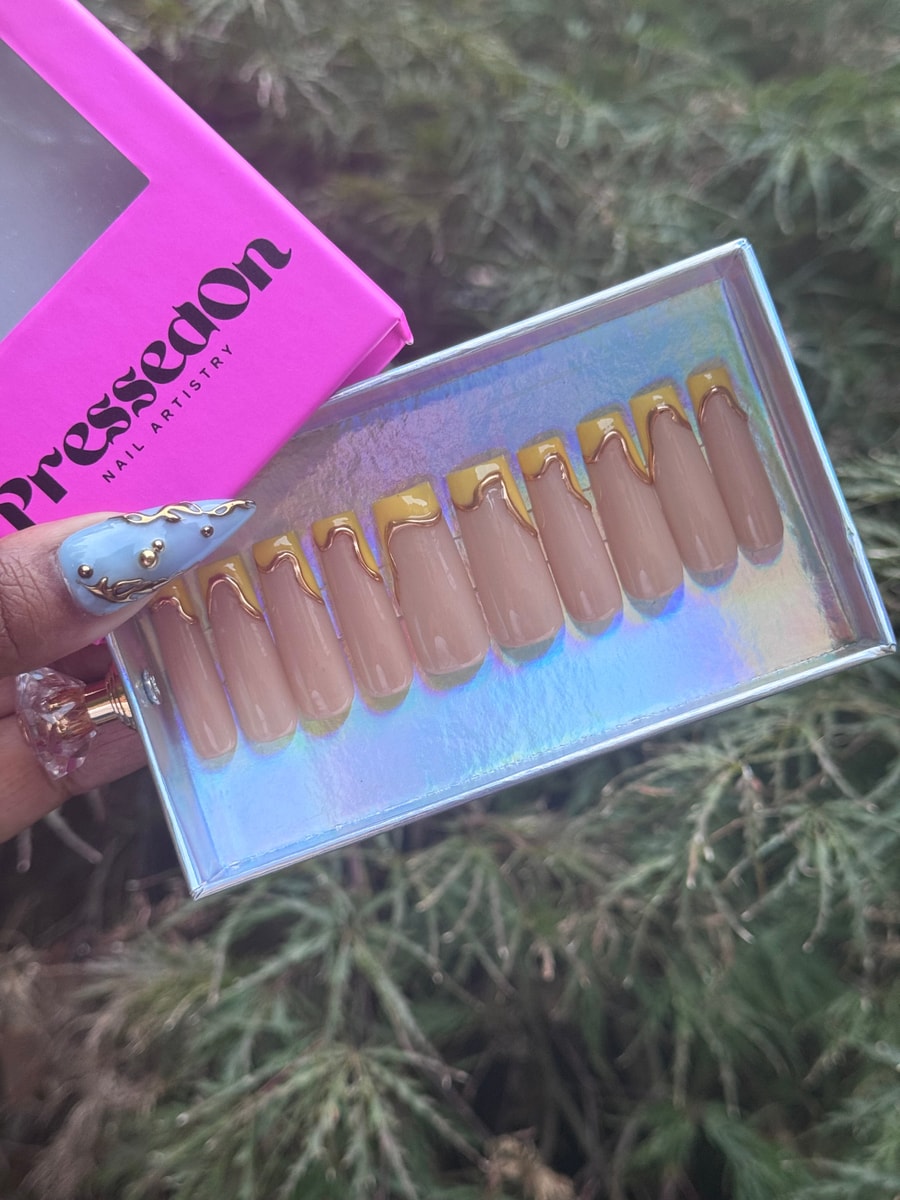 The image shows medium length nude press-on nails with dripping gold and yellow French tips, and one blue accent nail with gold details.