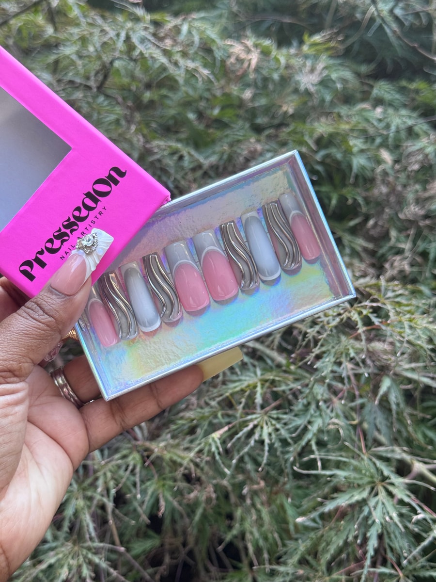 Medium length press-on nails are gray and pink, with some nails featuring white French tips and others having a wavy silver accent.