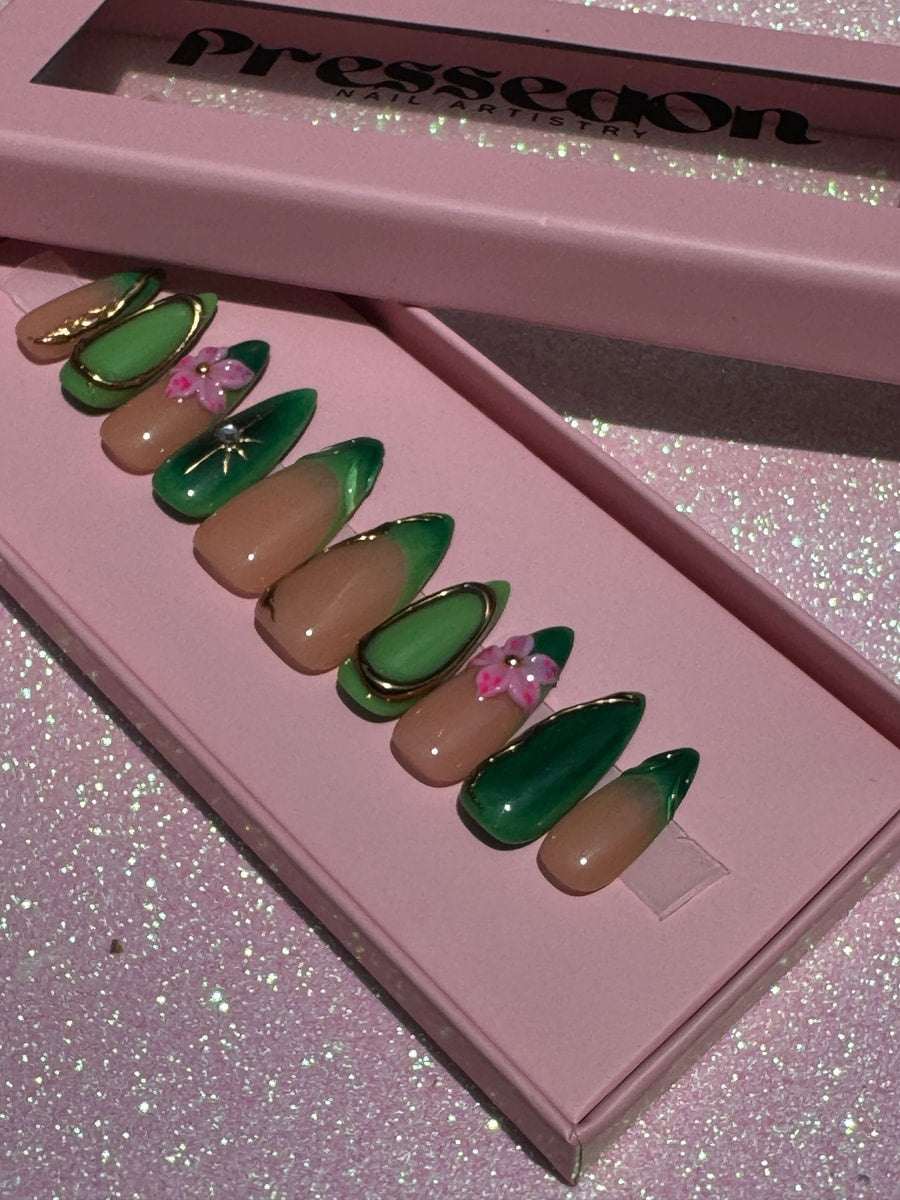 Short almond-shaped press-on nails in green and nude colors, with gold trim, a pink flower, and a starburst design.
