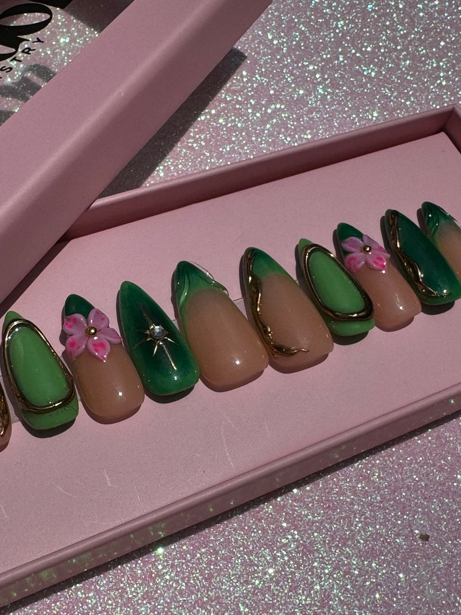 Almond-shaped press-on nails with a green tip, nude base, and gold accents. Some nails feature a pink flower charm or a gold starburst.