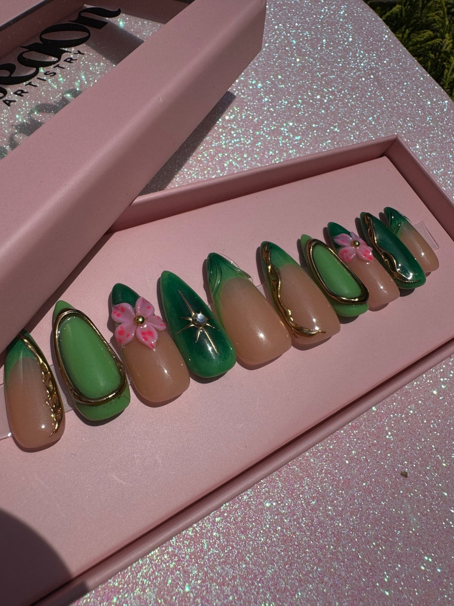 Short, almond-shaped press-on nails with a green and nude color scheme and gold accents. One nail features a 3D pink flower.