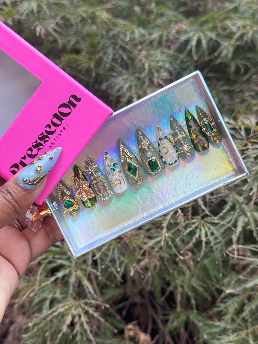 A set of short stiletto press-on nails in green, gold, and white, decorated with gems, crosses, floral designs, and miniature vintage frames.