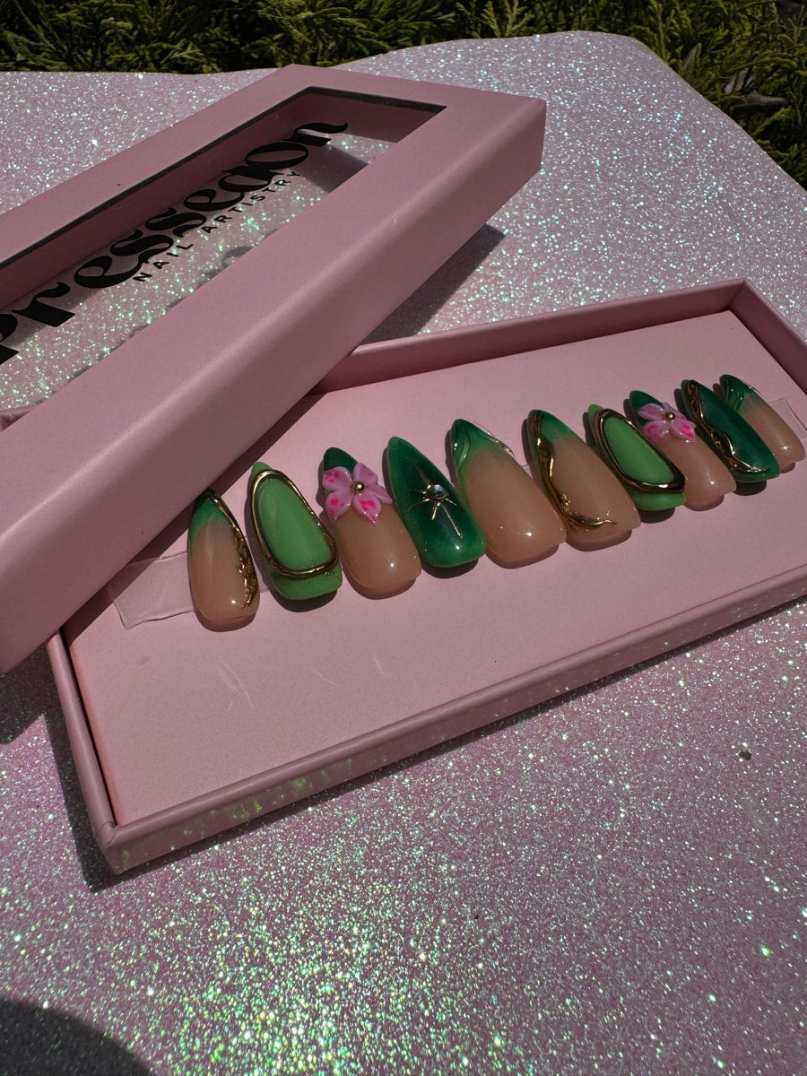 Set of nude and green short press-on nails. Designs include green tips, gold outlines, and a pink flower charm on one nail.