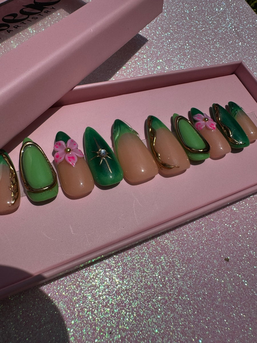 These are press-on nails with a combination of green and nude colors, accented by gold details. A 3D pink flower decorates one of the nails.