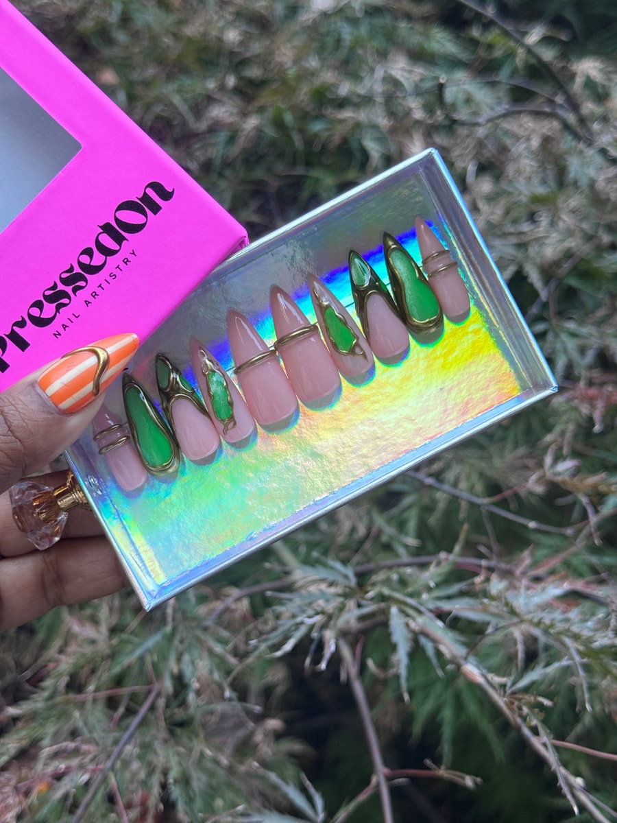 Medium-length press-on nails featuring a mix of nude and green designs with gold accents. Some nails have solid nude or green colors, while others have gold bands and gemstone details.