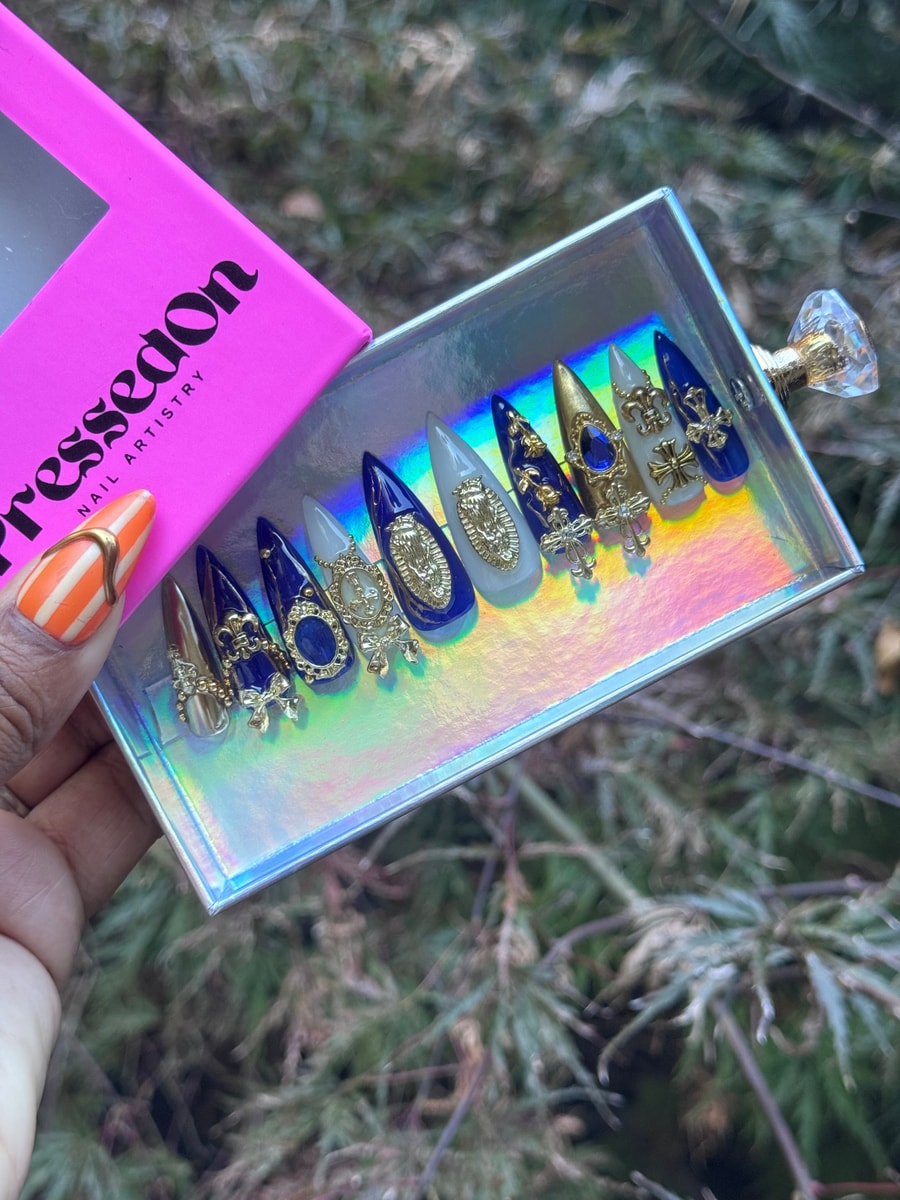 A set of medium-length press-on nails with a pointed shape, featuring a color palette of blue, white, and gold, and decorated with religious-themed gold charms.
