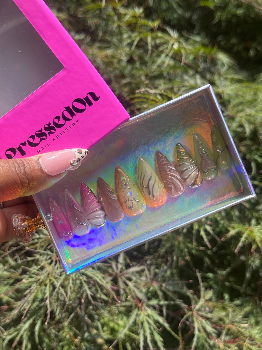 Medium length press-on nails feature butterfly wing designs in shades of pink, brown, and clear. Some have silver accents.