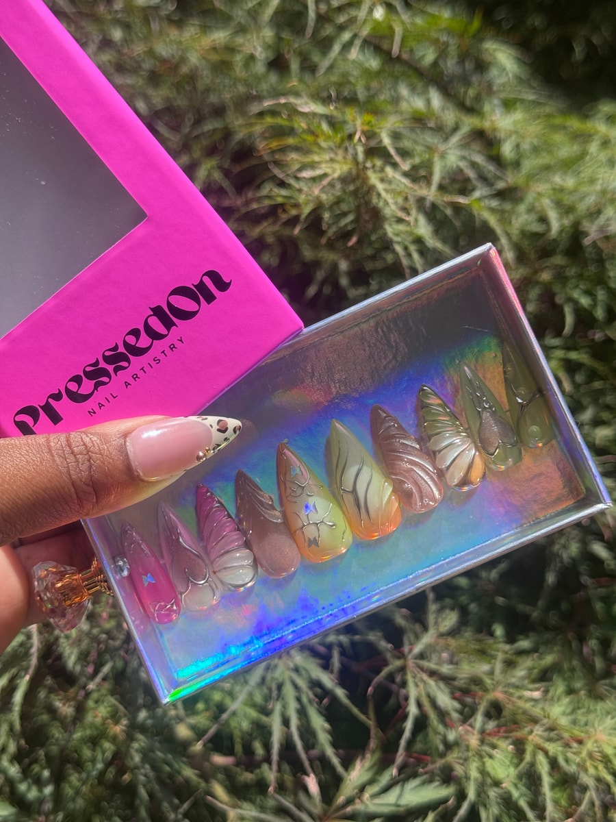 A set of medium-length press-on nails features a semi-sheer design with butterfly wings in pastel pink, brown, yellow, and green shades, creating an ethereal iced dream look.