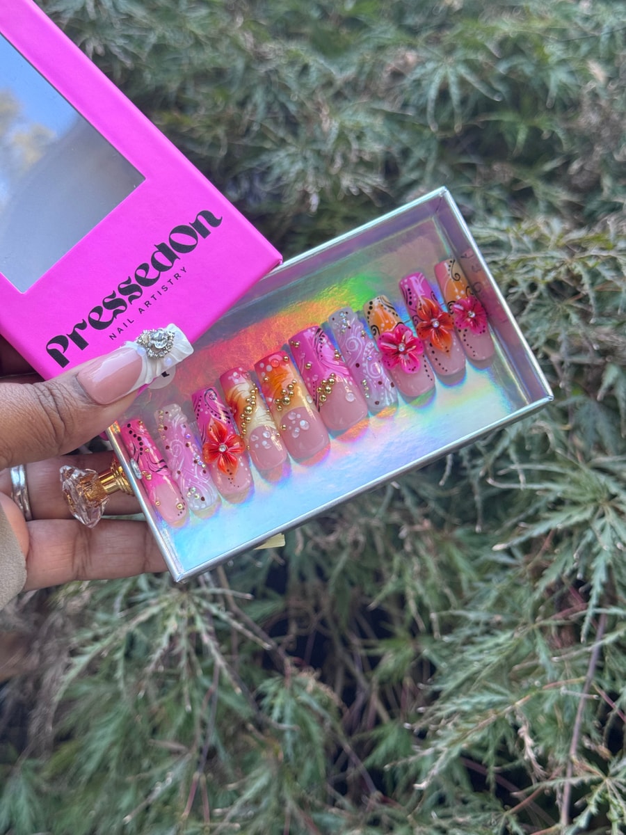 Medium length press-on nails feature a nude base with intricate floral designs in pink, orange, and white, accented with gold embellishments and gems.