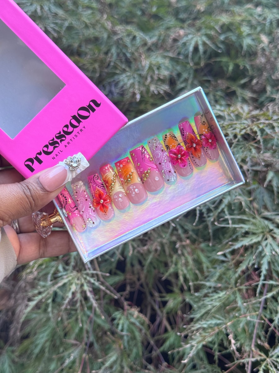 A set of medium-length island bloom press-on nails with a pink and orange gradient, white swirl designs, gold accents, and delicate 3D flower decorations.