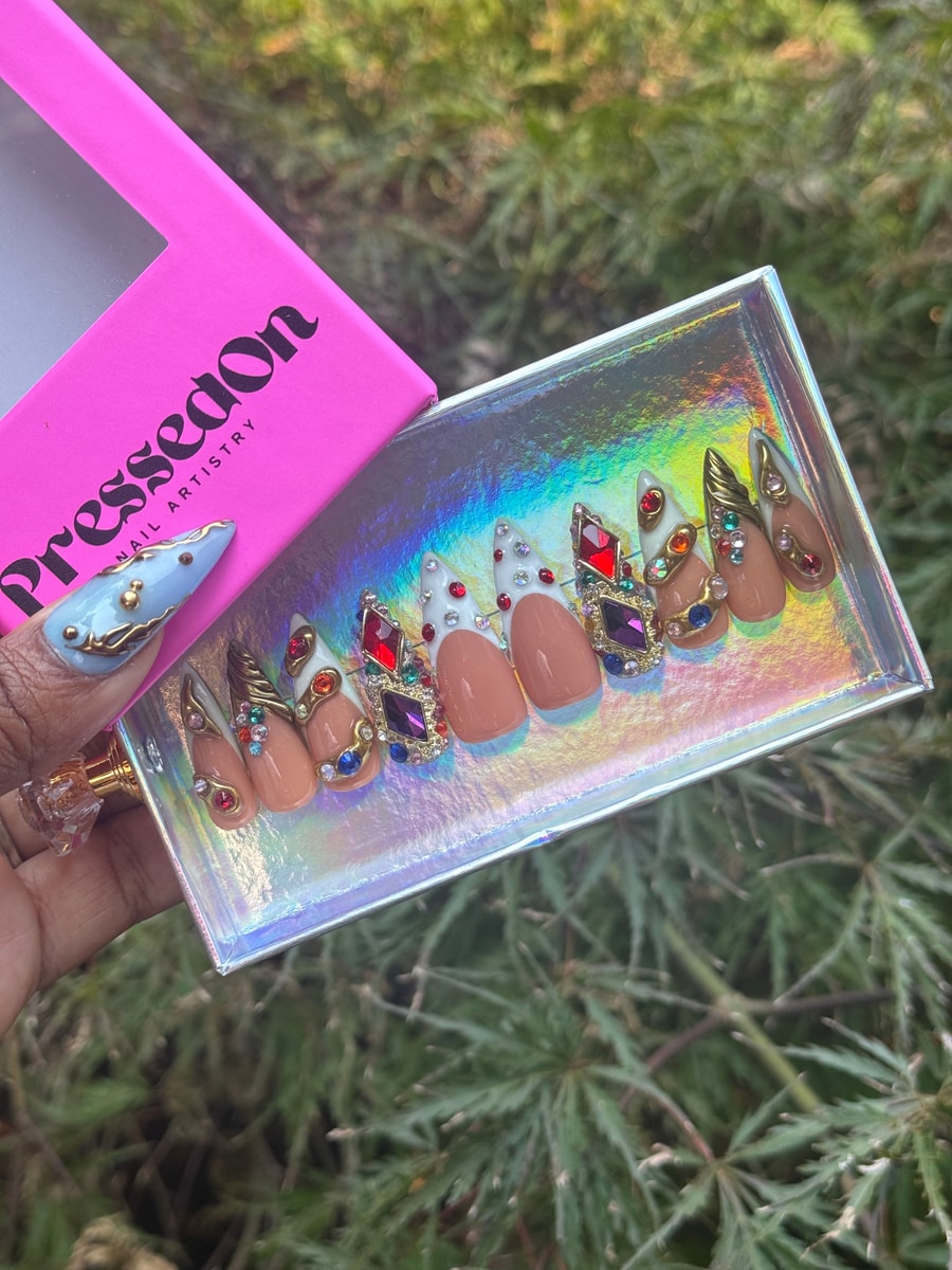 Short nude press-on nails with white tips are decorated with ornate jeweled embellishments. The gems are in shades of red, purple, blue, and gold.