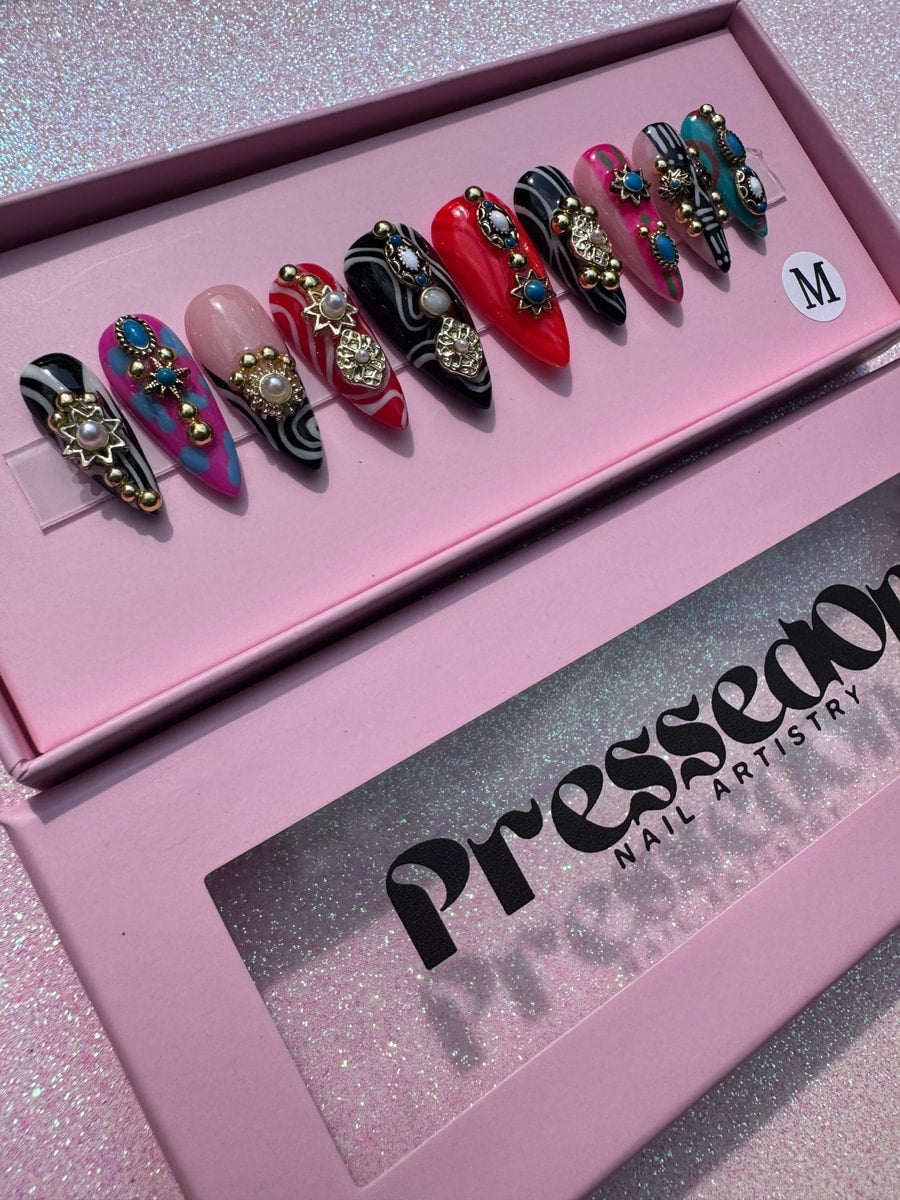 Super short press-on nails with a junk style featuring a variety of swirls, stone, and gold embellishments in black, pink, red and turquoise.