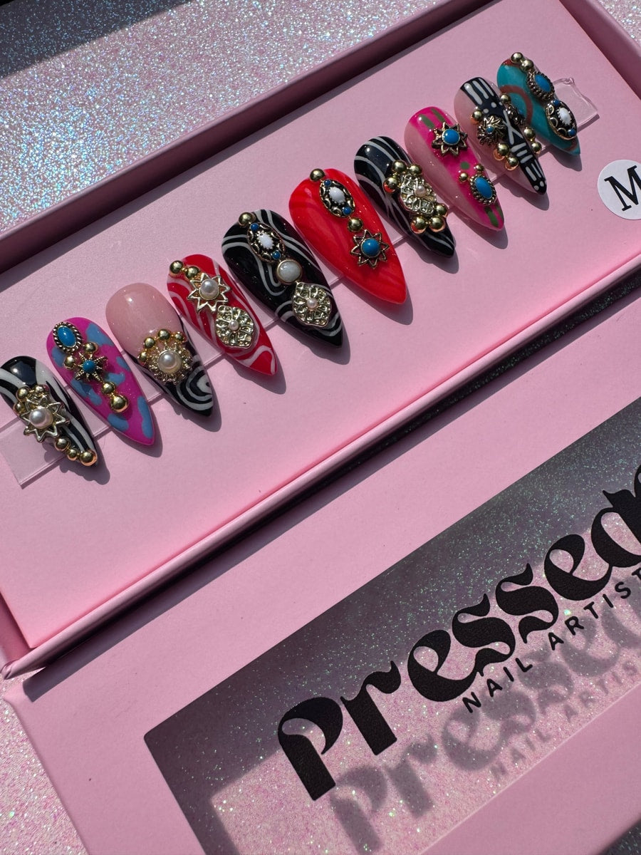 Set of short junk style press-on nails in a variety of colors and patterns, each decorated with ornate gold charms and gems.