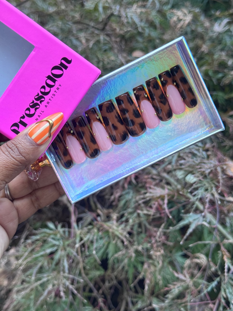 These are medium-length press-on nails with a leopard print design over a brown base, accented with light pink French tips and tiny gold studs at the base.