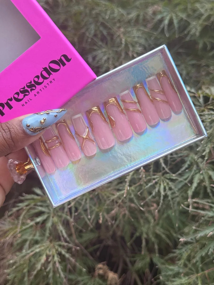 Medium length pink press-on nails with glossy finish and gold metallic line art. The designs include curved and criss-cross patterns on a classic square shape.